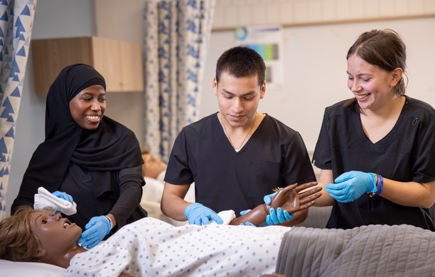 Three students gathered around a mannequin in a hospital simulation room, practicing medical procedures. 