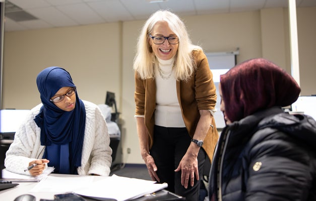 Three individuals collaborating around a desk in an office setting. One person, wearing a hijab, is seated and writing notes while another listens attentively. A third person, standing, smiles encouragingly while engaging with the group.