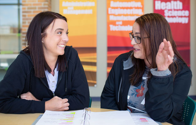 two students facing eachother talking at table and viewing a binder