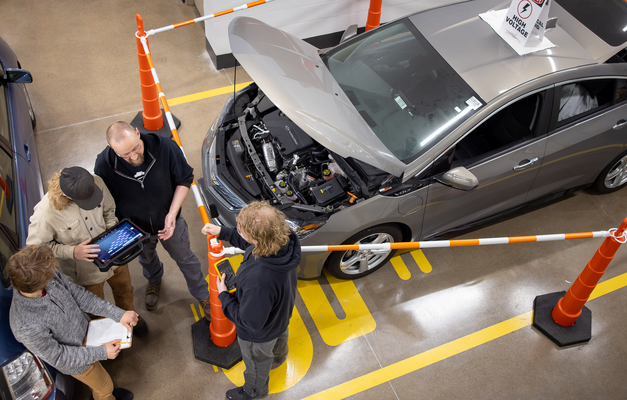 Top view of three individuals and an automotive technician discussing a car's engine in a workshop with safety cones around.