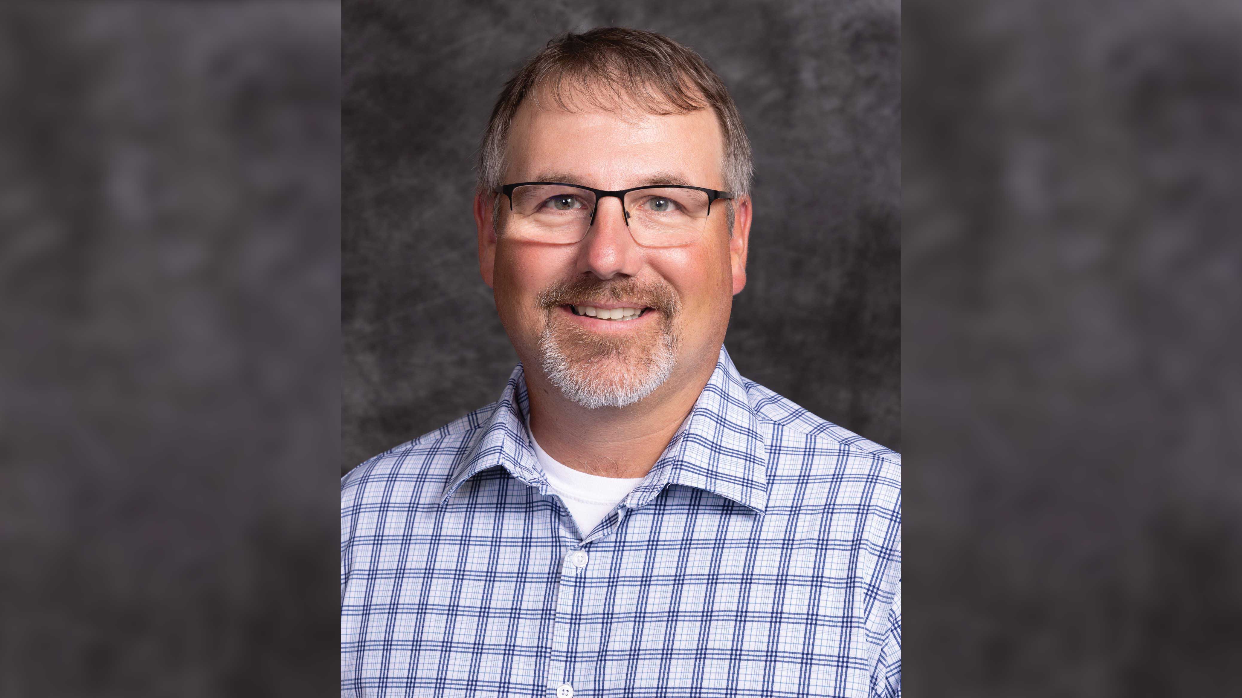 Portrait of an individual with glasses smiling, wearing a blue and white checkered shirt against a mottled gray background.