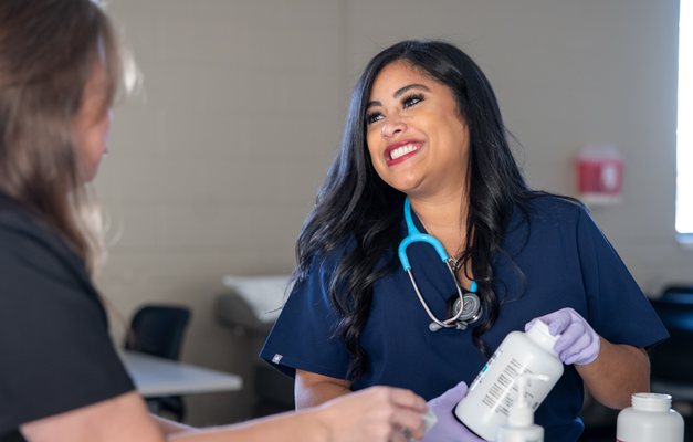 Two healthcare professionals in scrubs are smiling and engaging in conversation, one holding a medication bottle in a clinical setting.